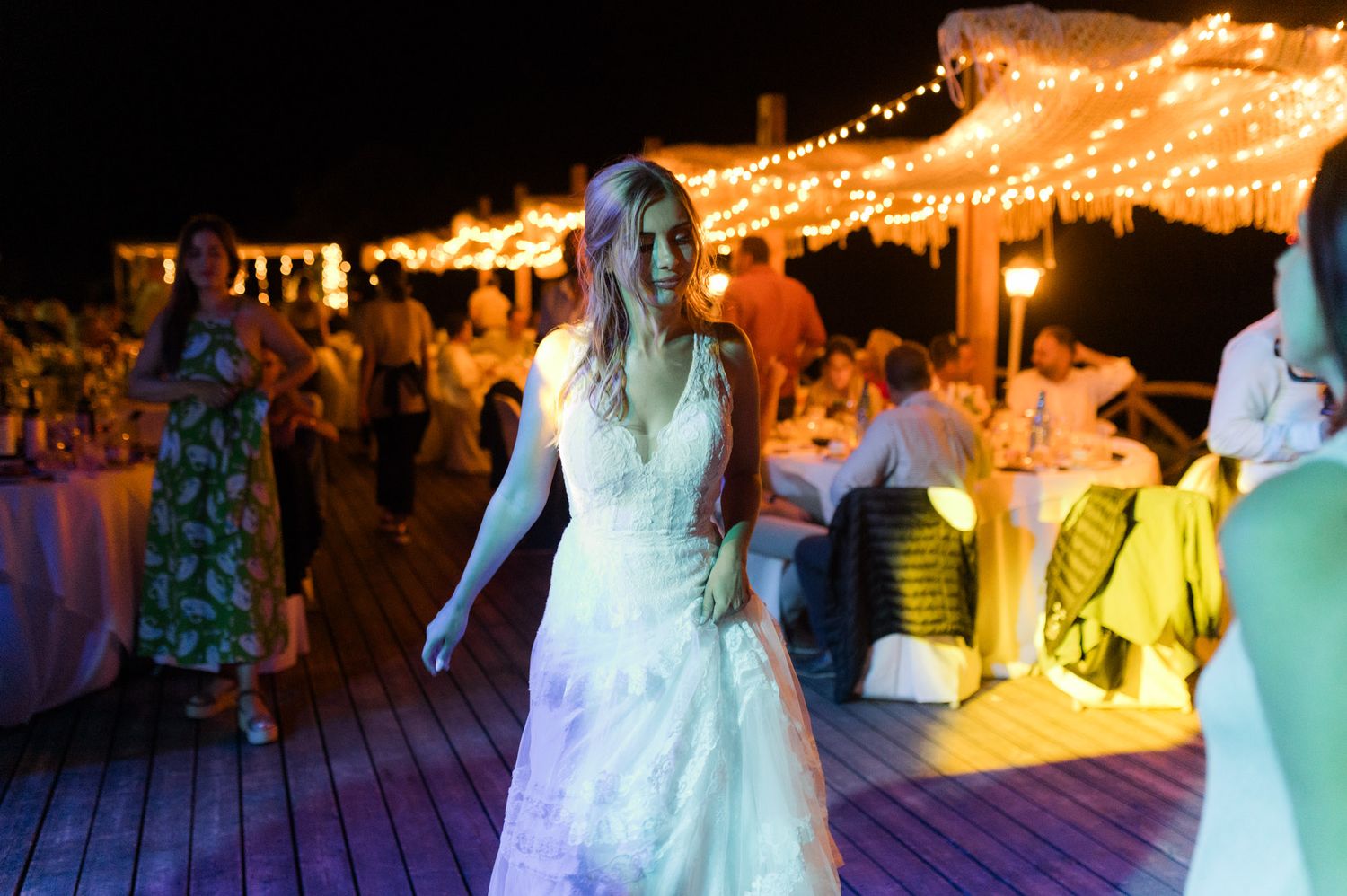 Bride in white lace wedding dress dancing at outdoor night reception with string lights.