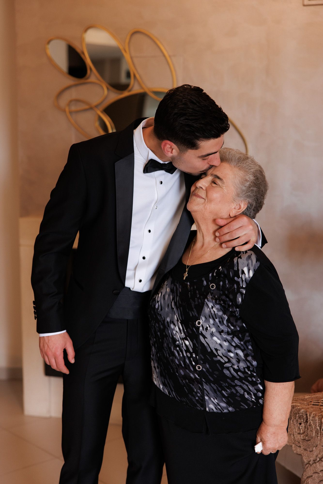 Groom in black tuxedo kissing elderly woman on cheek in elegant indoor setting.
