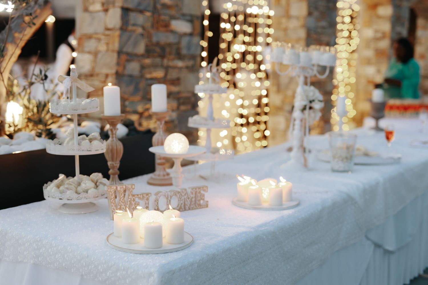 Elegant white wedding dessert table with candles, tiered stands, and warm fairy lights against a stone wall backdrop.