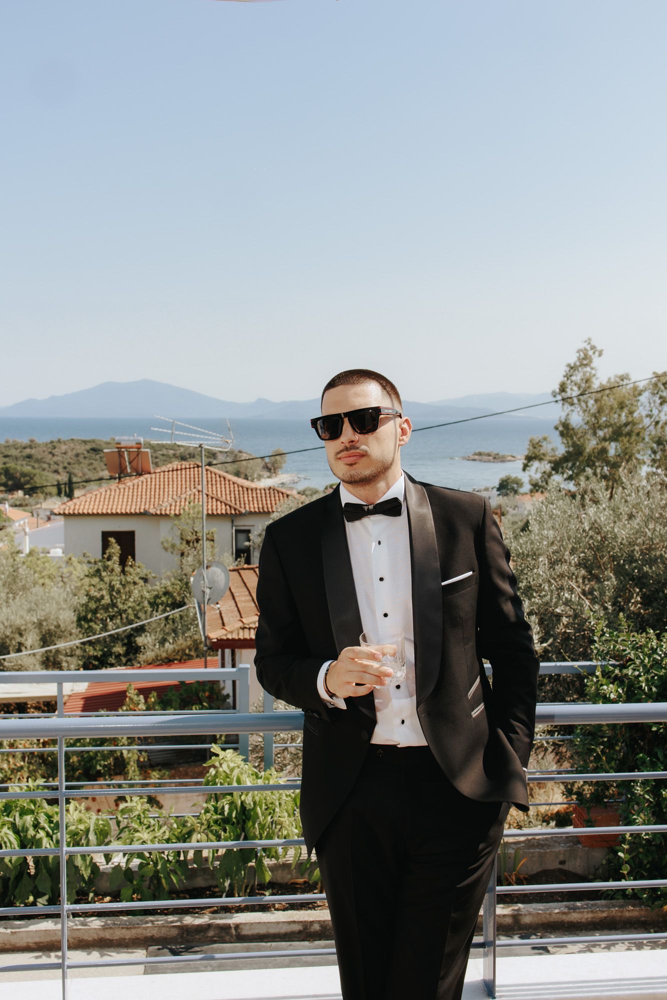 Groom in black tuxedo and sunglasses holding champagne on balcony with Mediterranean coastal view.