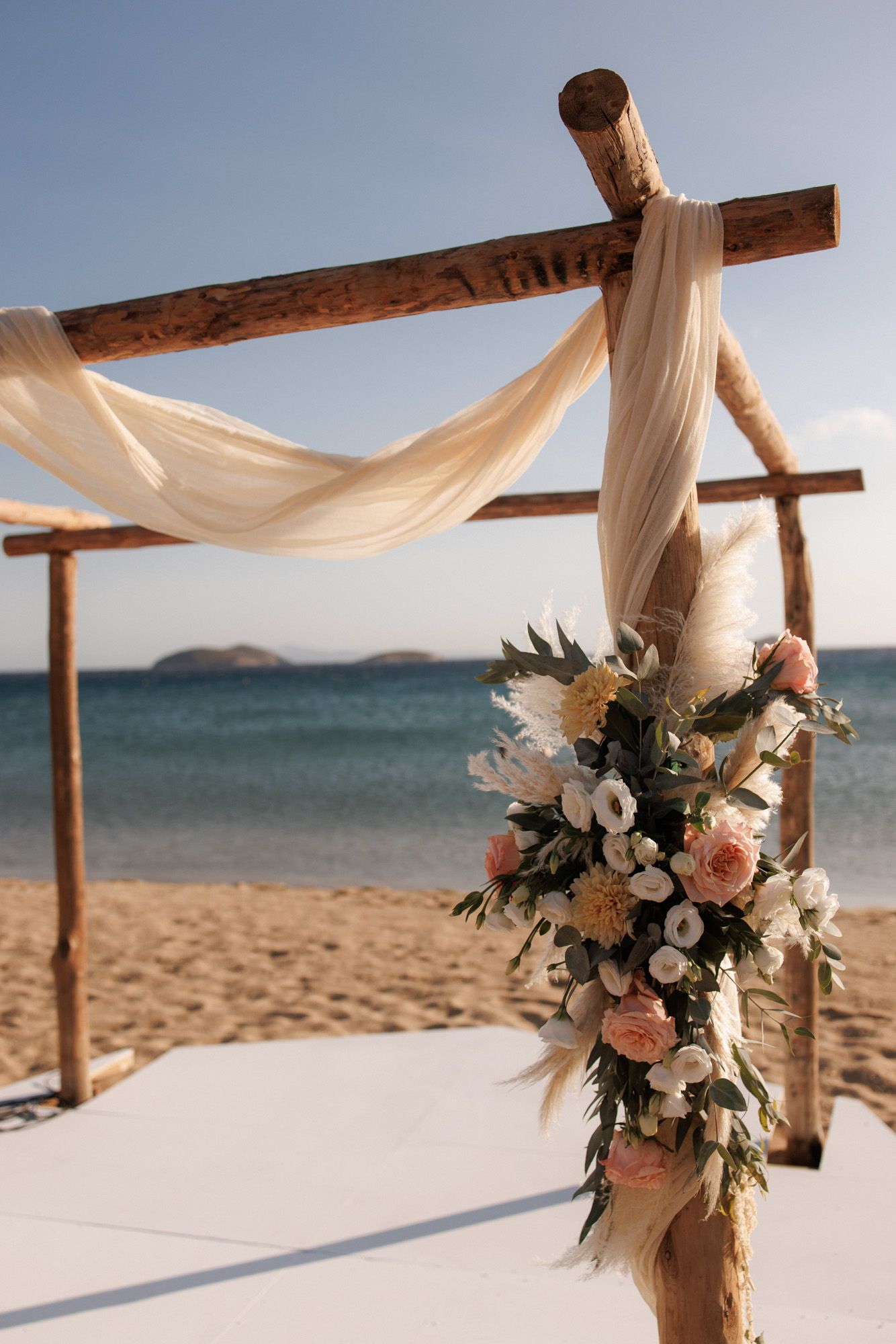 Rustic wooden beach wedding arch adorned with pink roses and pampas grass flowers.
