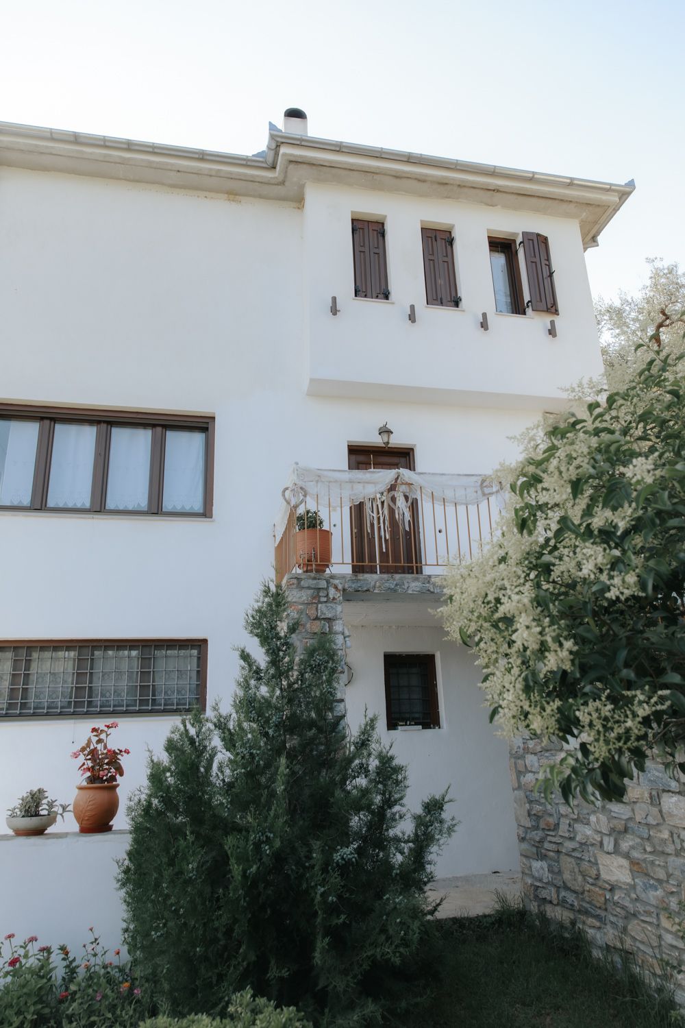 White two-story Mediterranean villa with balcony, wooden shutters, terracotta pots, and lush green garden shrubs.