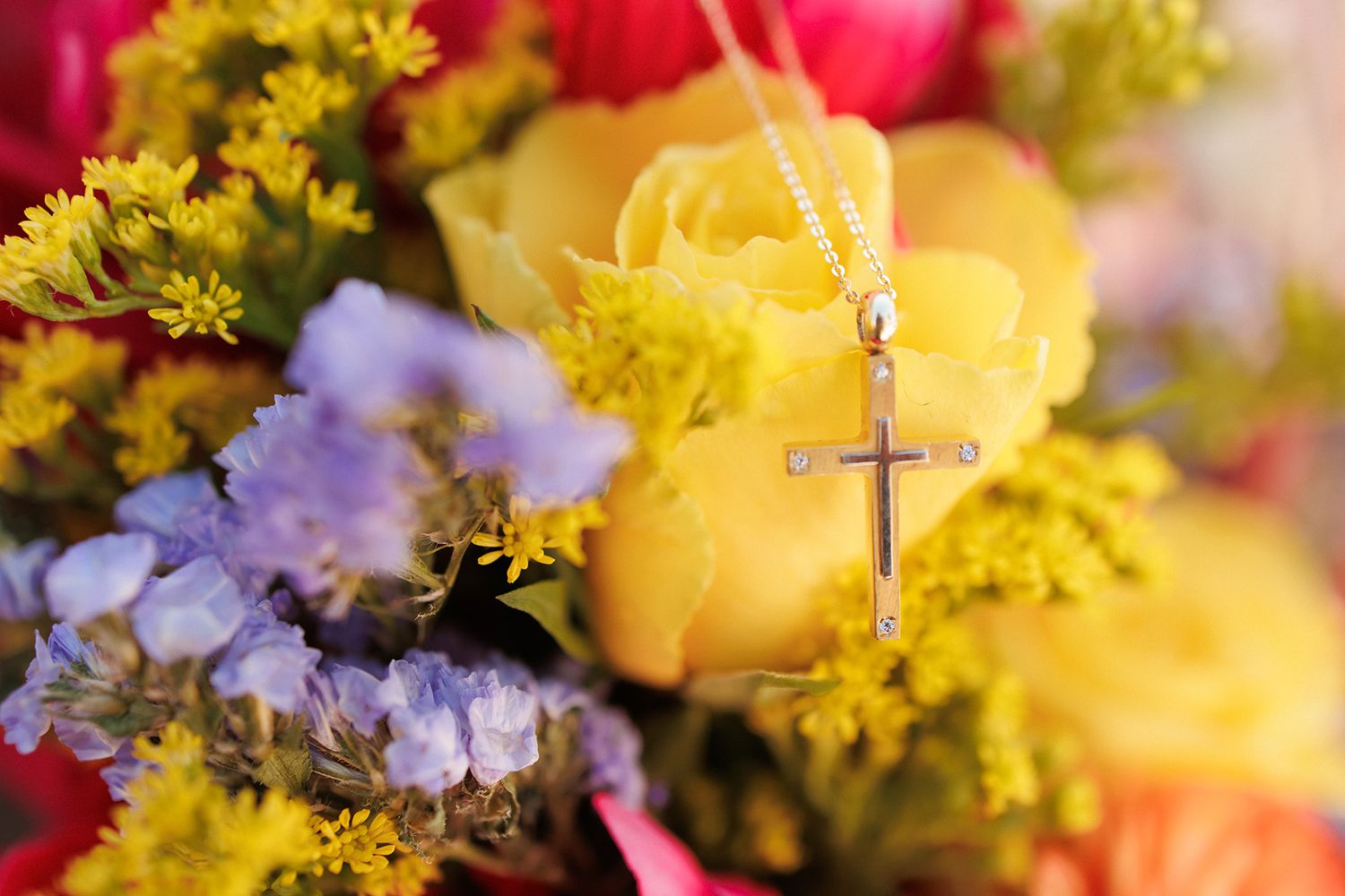 A silver cross necklace resting on a vibrant bouquet of yellow roses, purple flowers, and colorful spring blooms.