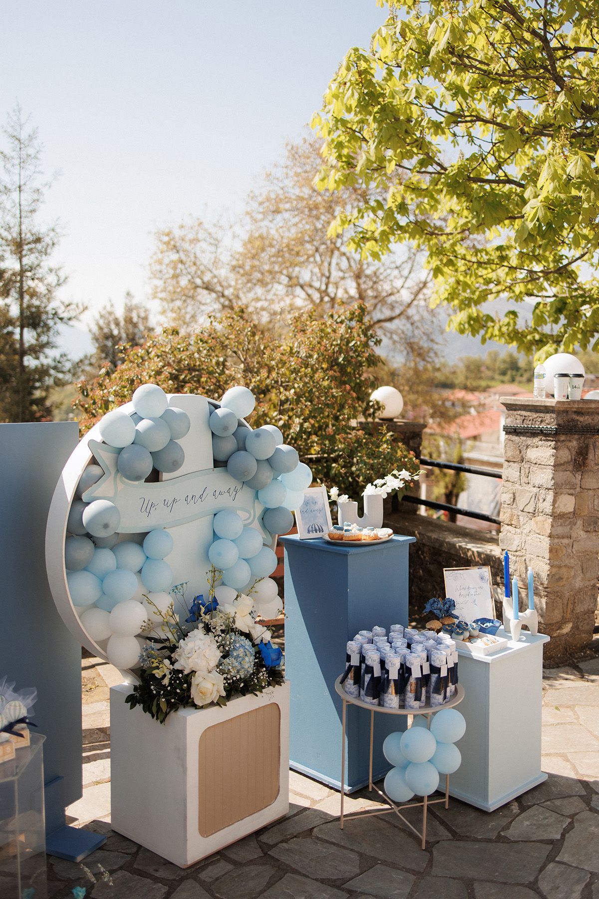 Outdoor baby shower setup with light blue balloon arch, floral arrangements, and elegant display tables under sunny skies.