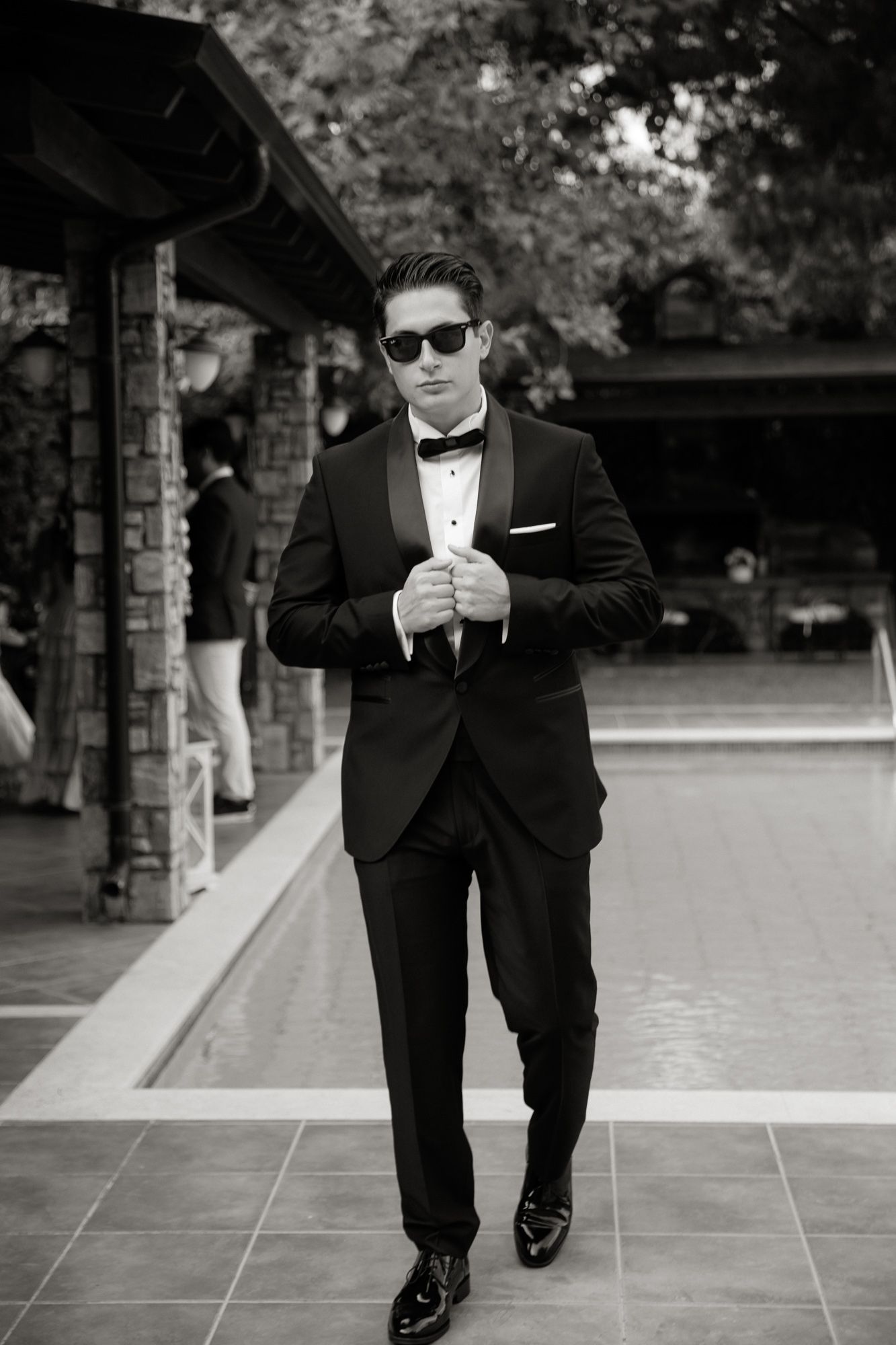 Dapper groom in black tuxedo and sunglasses posing confidently outdoors in a black and white photo.