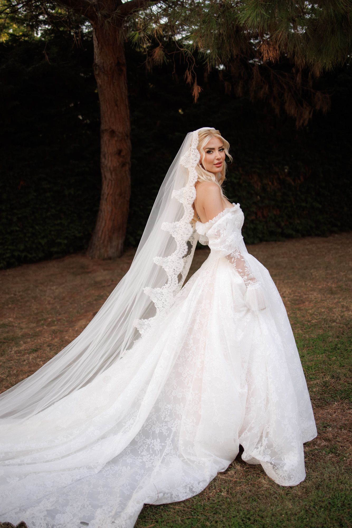 Stunning blonde bride in intricate lace off-shoulder ball gown with long cathedral veil turns gracefully in garden.