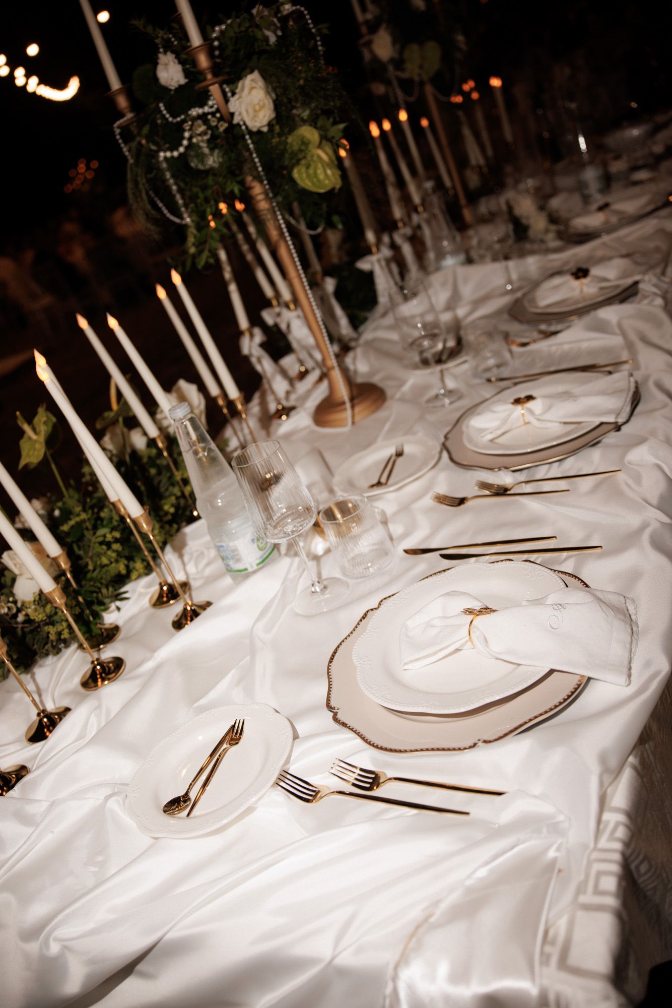 Elegant white wedding reception table set with gold cutlery, candles, and floral centerpieces.