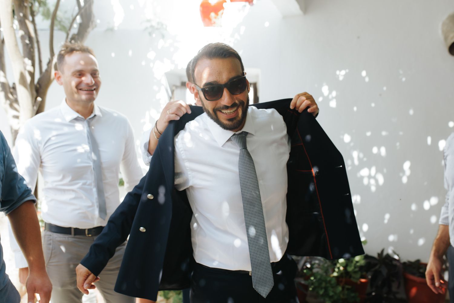 Cheerful groom in sunglasses putting on suit jacket surrounded by smiling groomsmen outdoors.