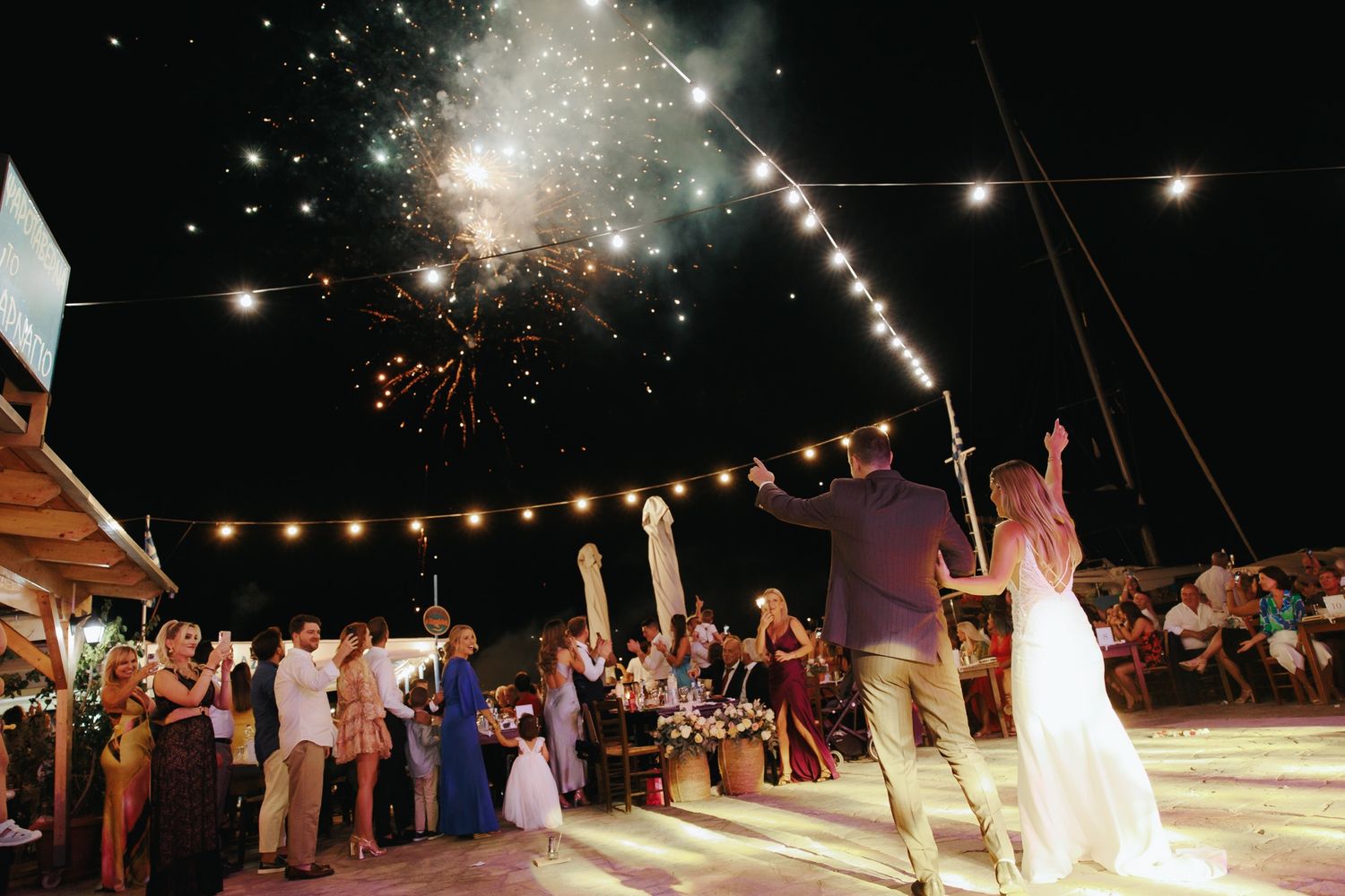 Newlyweds celebrate with raised hands as fireworks explode above outdoor wedding reception at night.