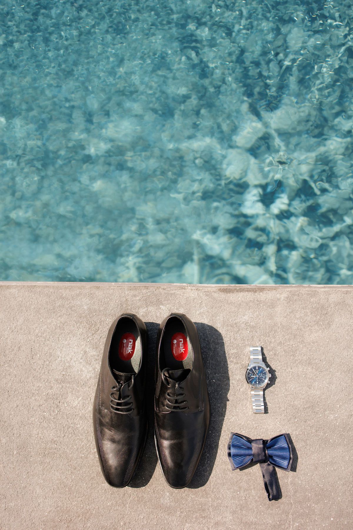 Black leather dress shoes, silver watch, and blue bow tie arranged by a poolside.