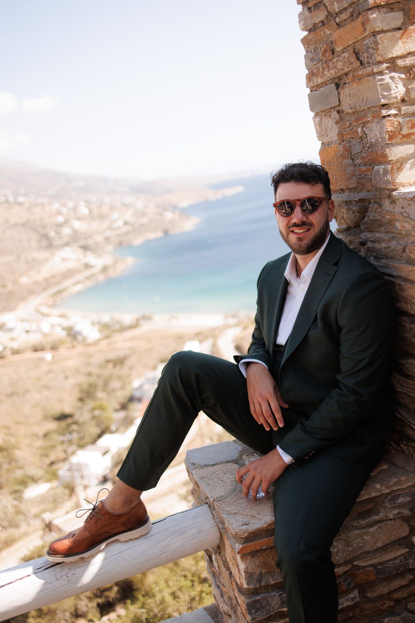 Stylish groom in dark green suit and sunglasses sitting on stone ledge with Mediterranean coastal view.