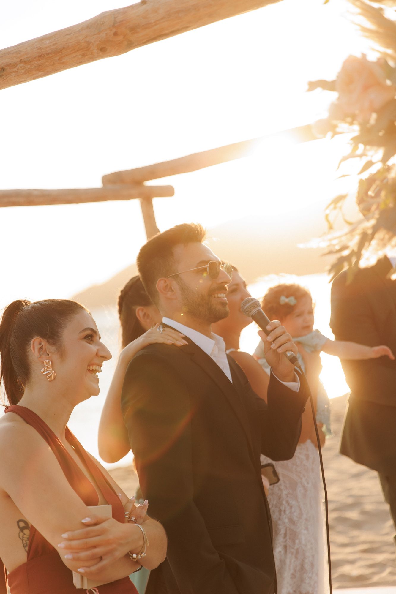 A man in a black suit speaks into a microphone at a golden hour beach wedding ceremony.