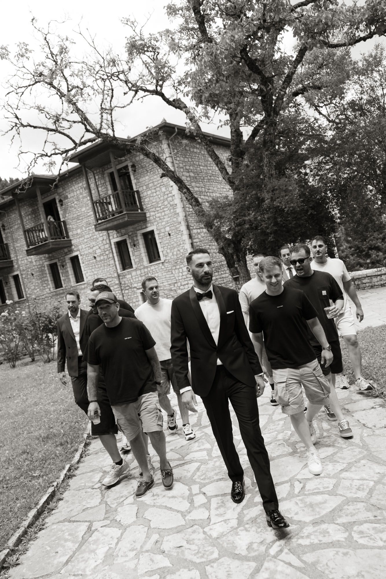 Black and white photo of dapper groom leading groomsmen walking outdoors near stone building.