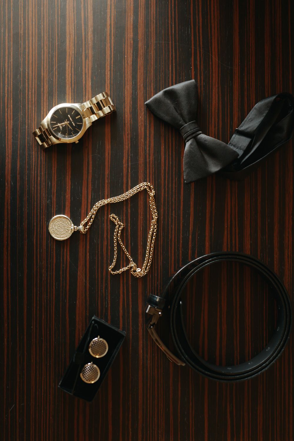 Gold watch, bow tie, belt, cufflinks, and chain necklace arranged on dark wood surface for a groom's wedding accessories flatlay.