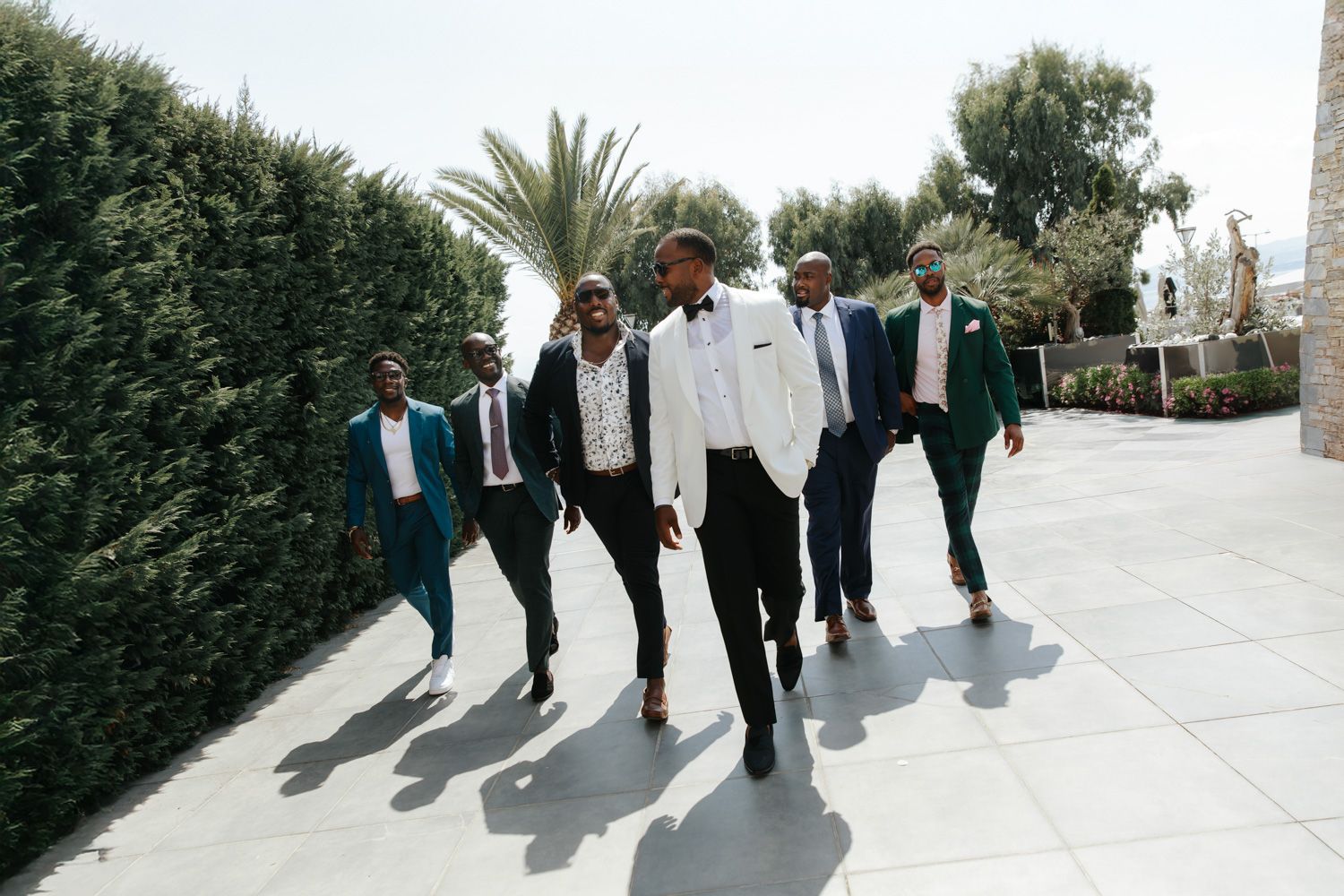 Groomsmen in teal and white suits walking confidently outdoors near hedges and palm trees on a sunny wedding day.