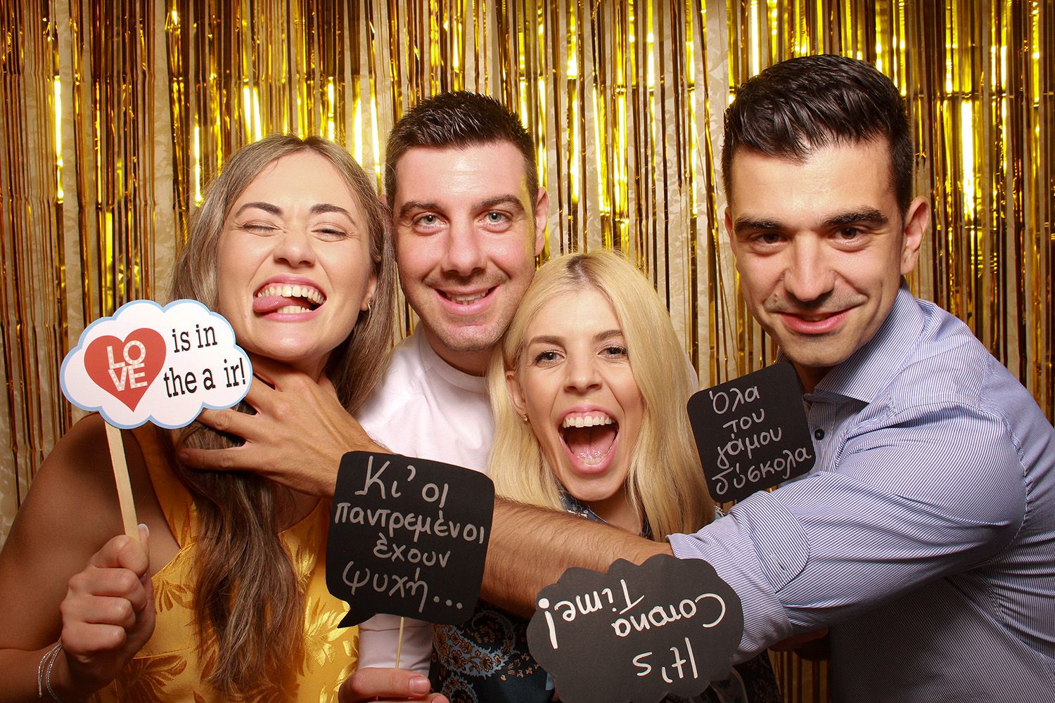 Four friends smiling and holding fun photo booth props at a gold glitter party celebration.