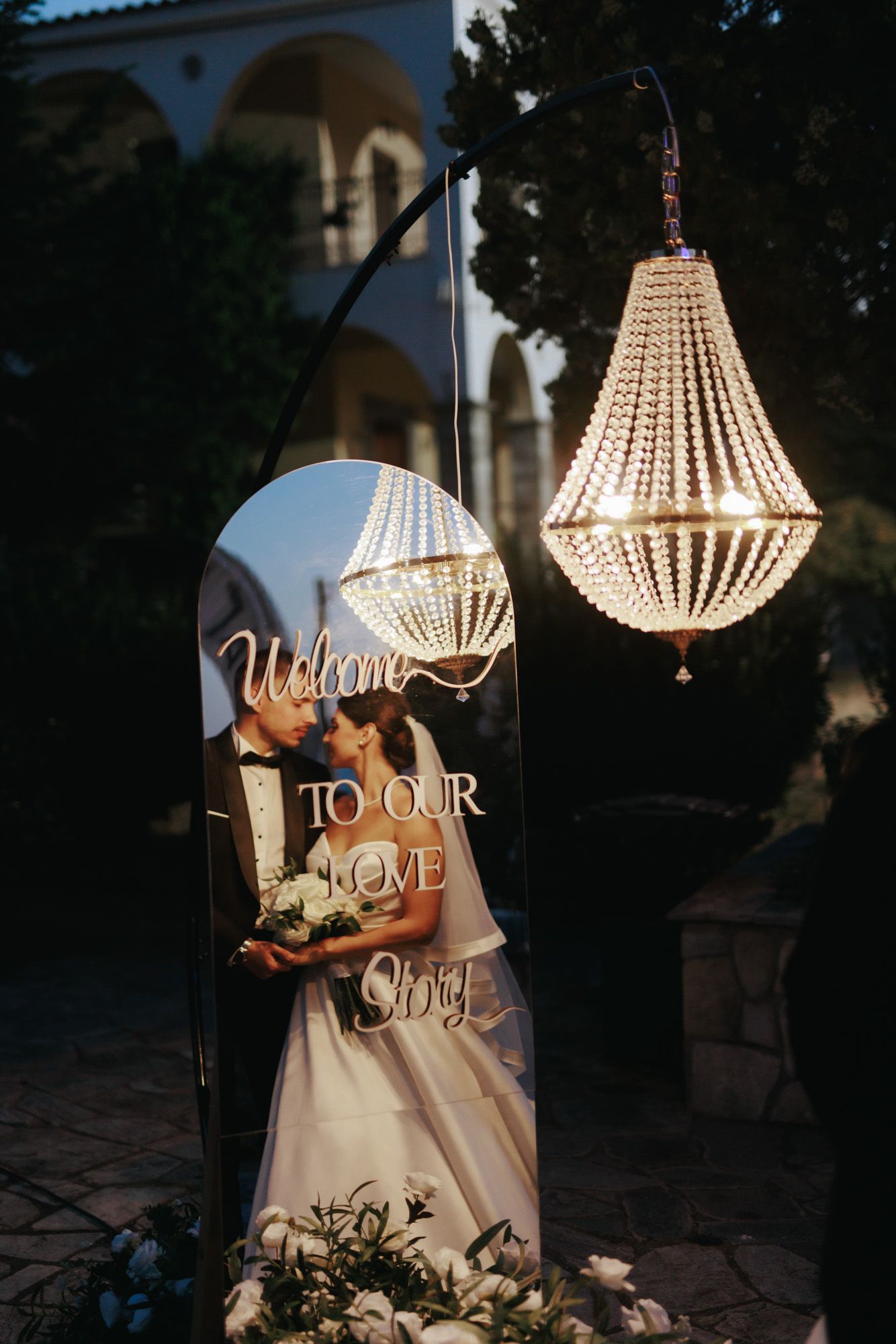 Elegant wedding welcome sign with crystal chandelier and couple photo displayed at outdoor evening ceremony.