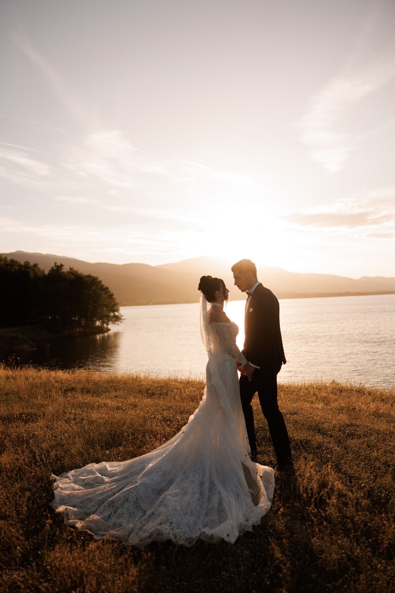 Bride and groom share a romantic moment at sunset by a lake, with mountains in the background and a flowing lace wedding gown.
