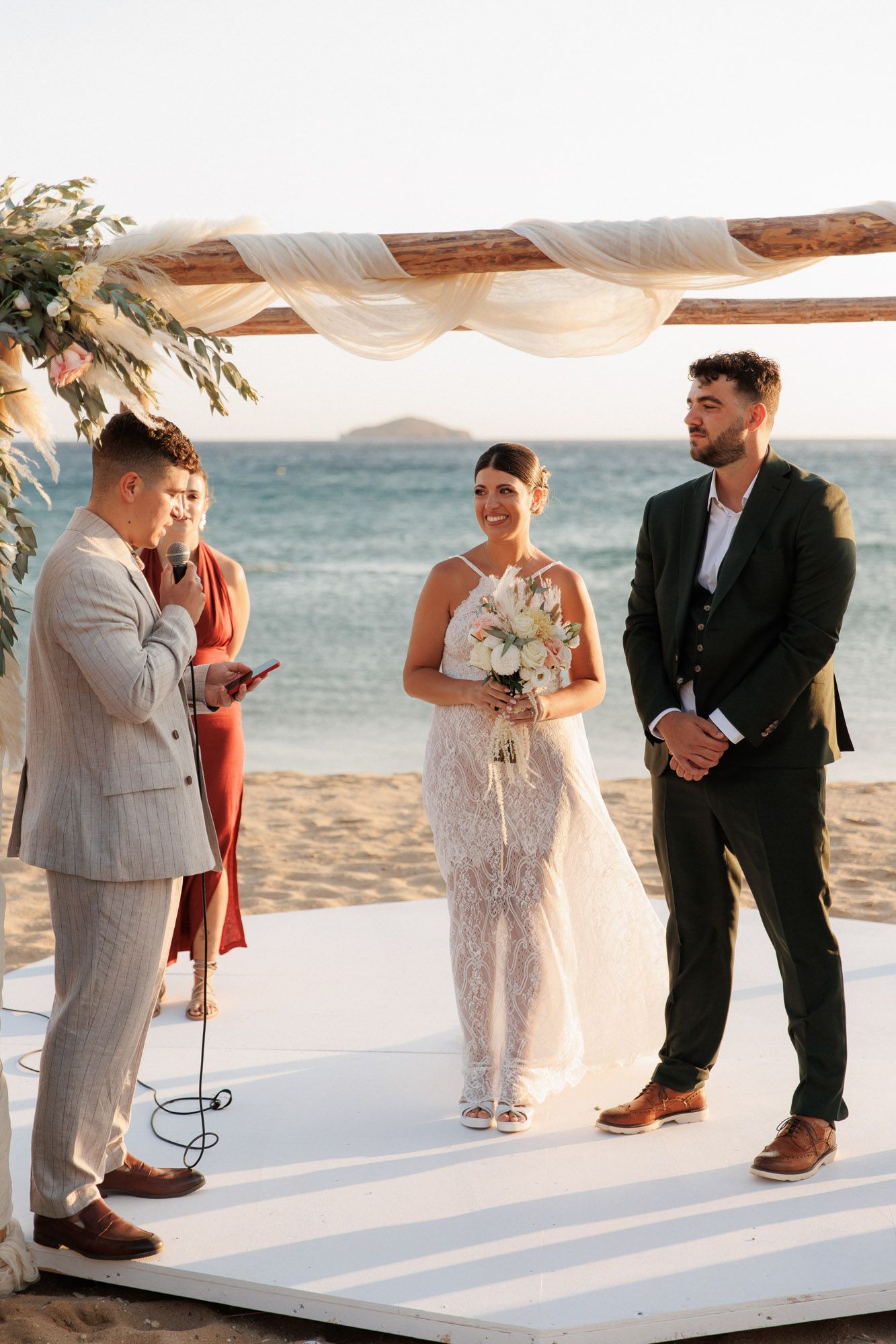Bride holding bouquet and groom stand at beach wedding altar while guest speaks at microphone.