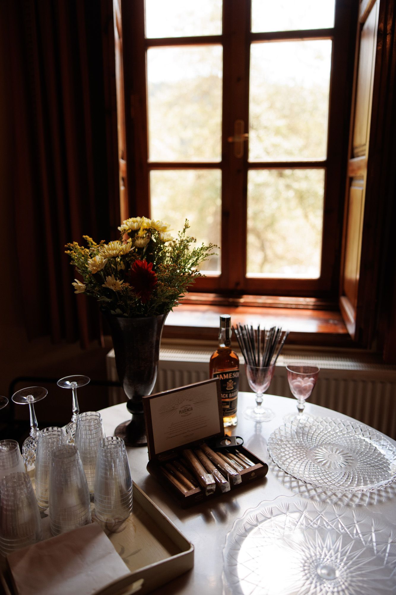 Elegant table setup with cigars, whiskey, crystal glasses, and flowers by a wooden window.