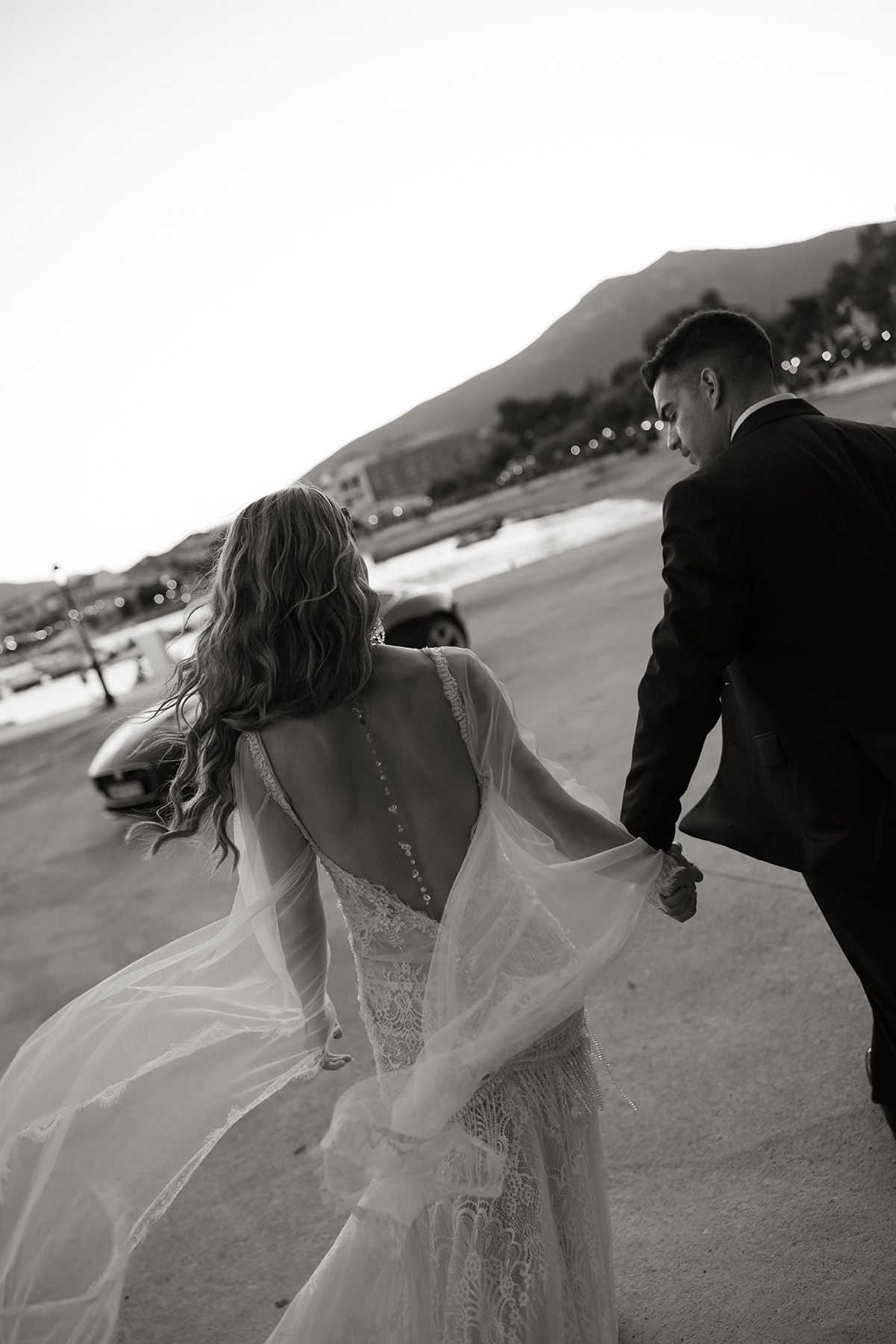 Black and white photo of bride and groom walking hand in hand at dusk, brides open-back gown trailing behind.