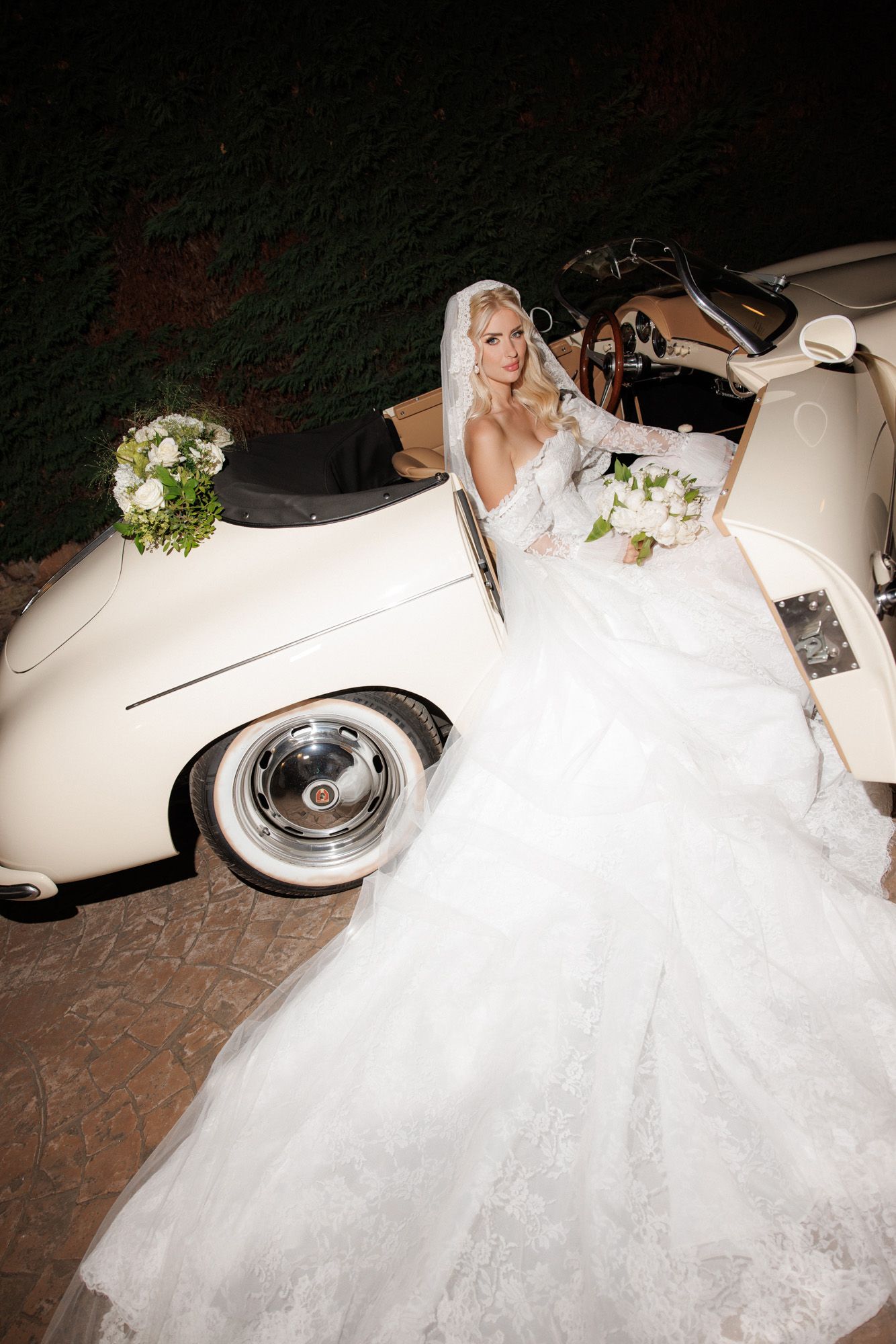 Beautiful bride in layered lace wedding dress standing beside vintage cream convertible decorated with white flowers.