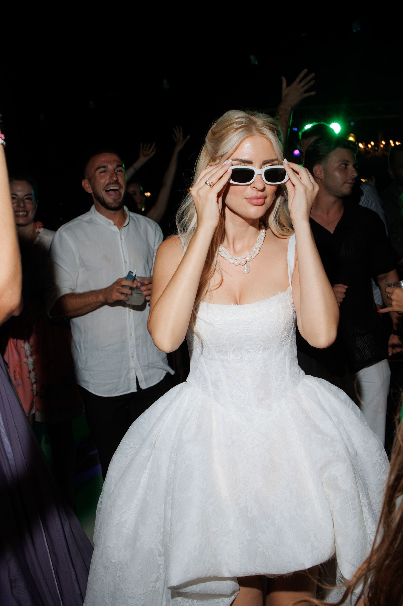 Blonde bride in white dress adjusting white sunglasses while dancing at wedding reception.