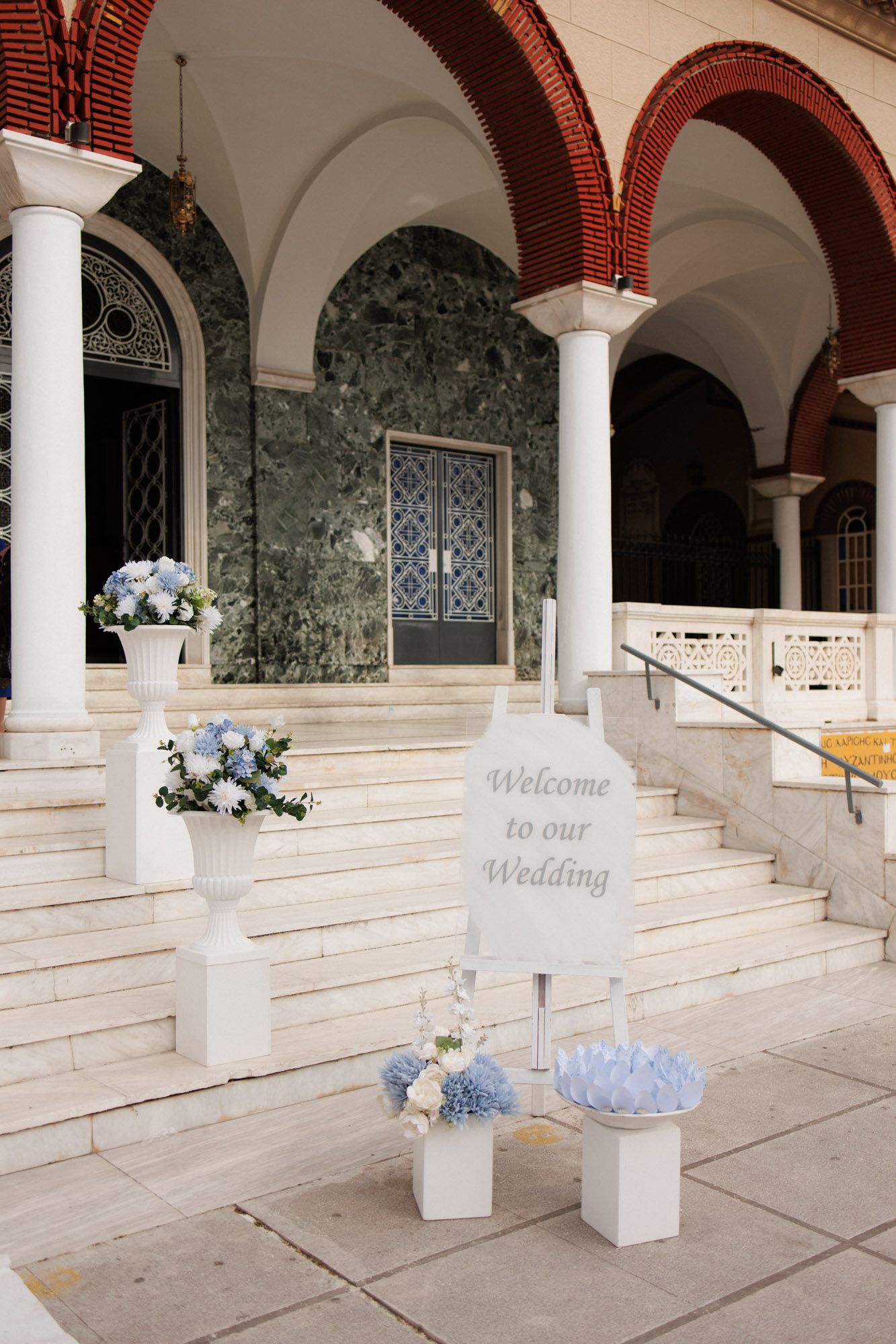 Elegant wedding venue entrance with white pedestal flower arrangements, blue hydrangeas, and a Welcome to Our Wedding sign.