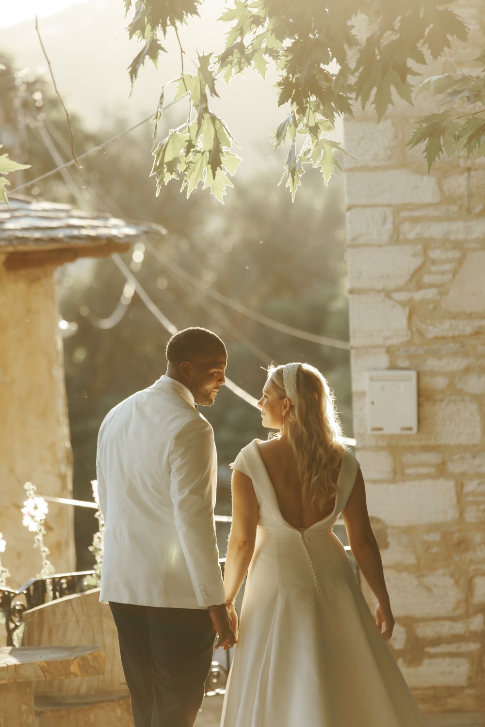 A bride and groom share a romantic moment in golden sunlight beside a stone building, surrounded by lush greenery.