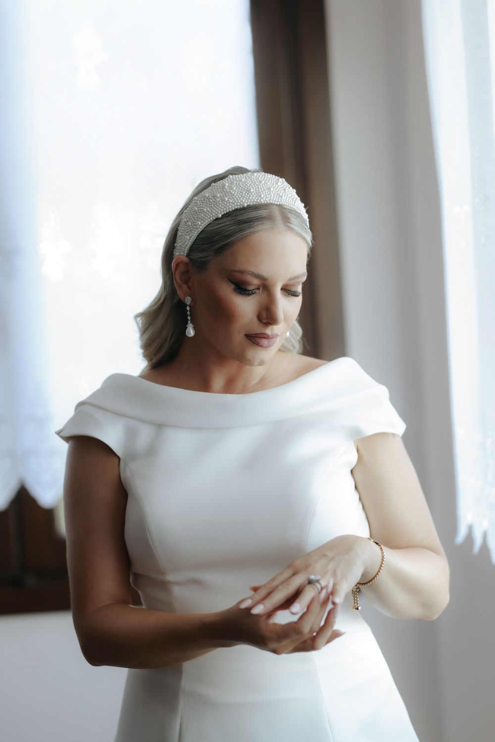 Elegant bride in white cap-sleeve gown and pearl headband gazes at her ring near a bright window.