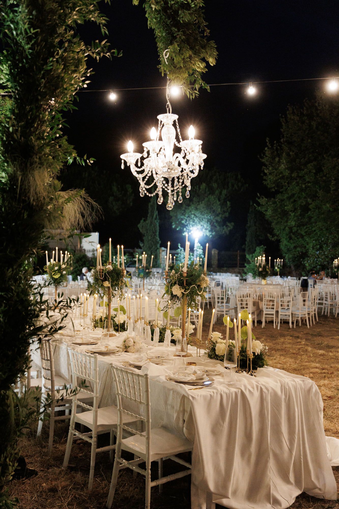Romantic outdoor wedding reception with white chiavari chairs, crystal chandelier, candlelit tables and string lights at night.