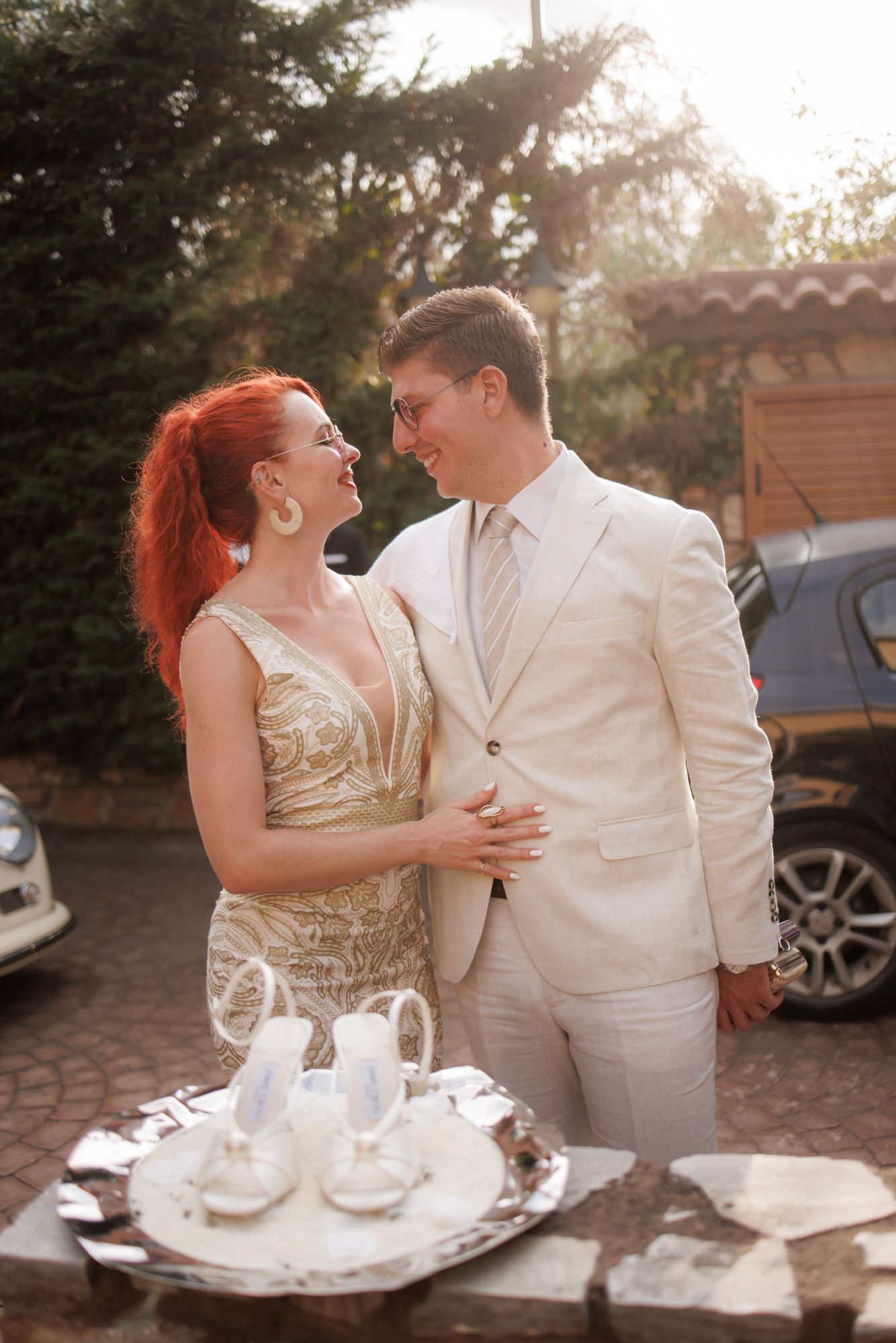Redhead woman in gold dress embraces smiling groom in white suit outdoors in golden sunlight.