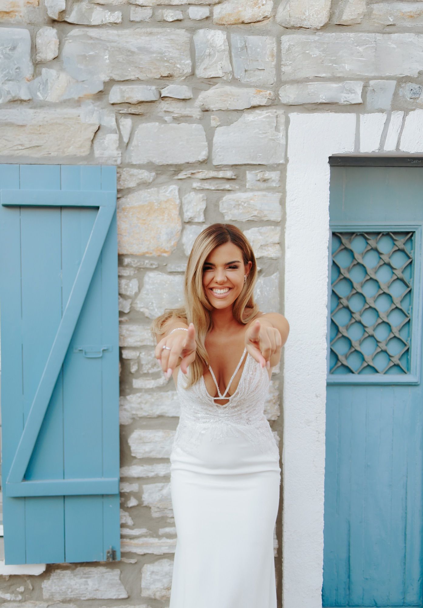 Joyful bride in white lace dress reaching toward camera in front of stone wall with blue shutters.