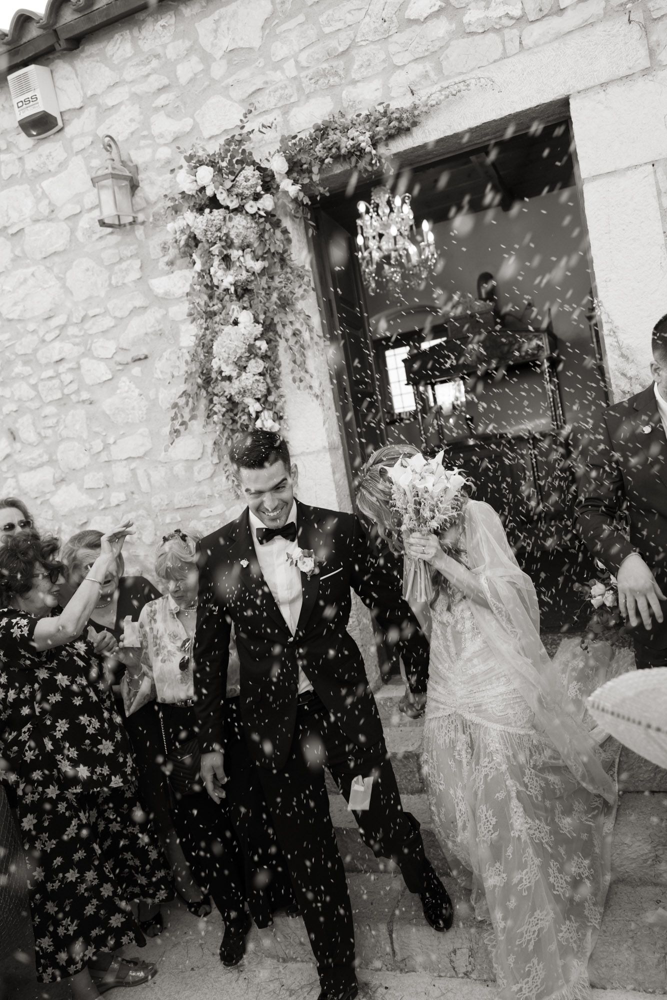 Black and white image of bride and groom walking through rice shower outside rustic stone church.
