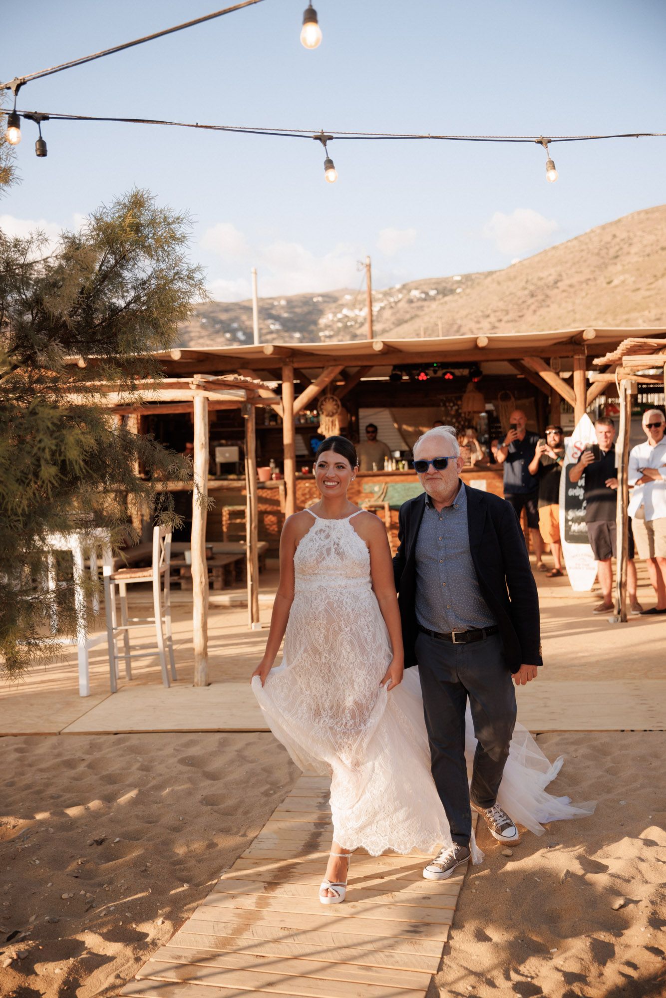Bride in lace gown walks with older gentleman along sandy beach under string lights at sunset.
