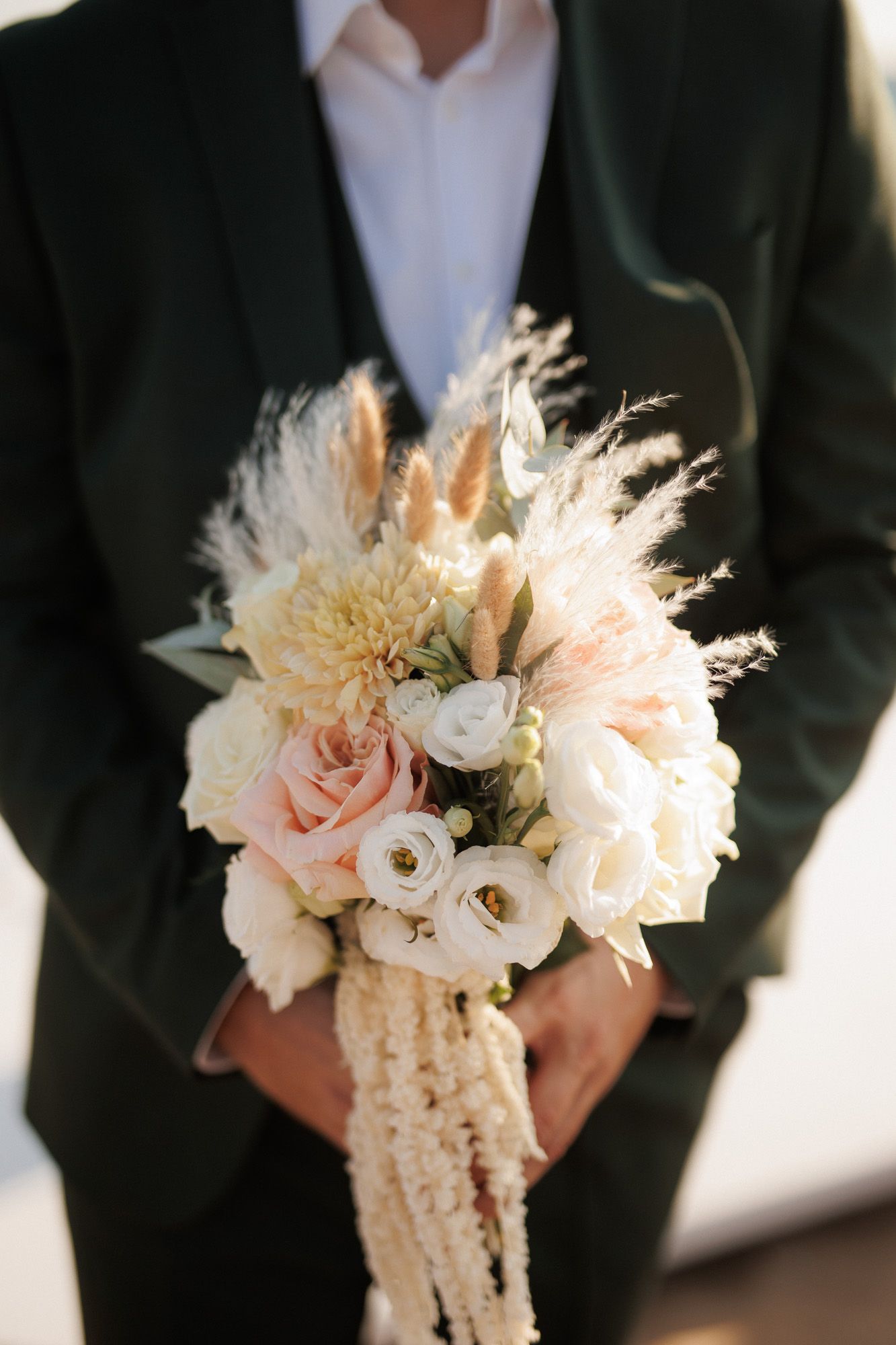 Groom in dark suit holding beautiful bridal bouquet of roses and pampas grass.