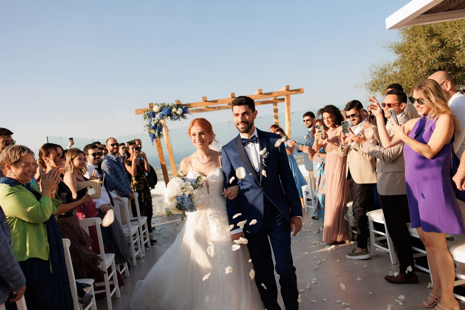 Newlyweds walk down aisle as guests toss petals at outdoor Santorini wedding ceremony.