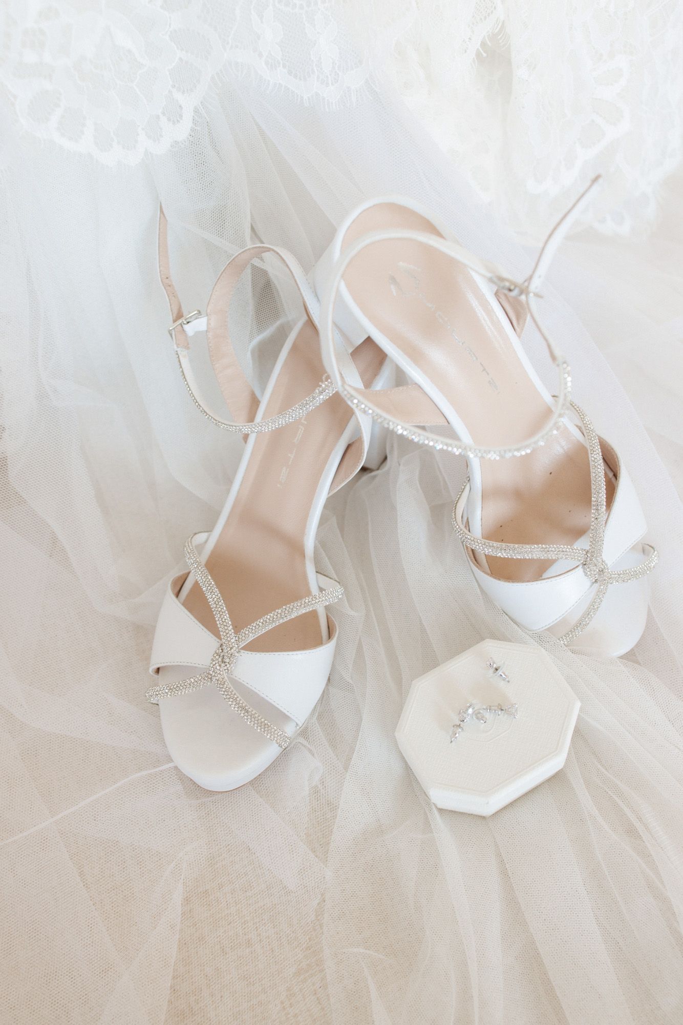 White strappy bridal heels with crystal details resting on veil next to jewelry box.