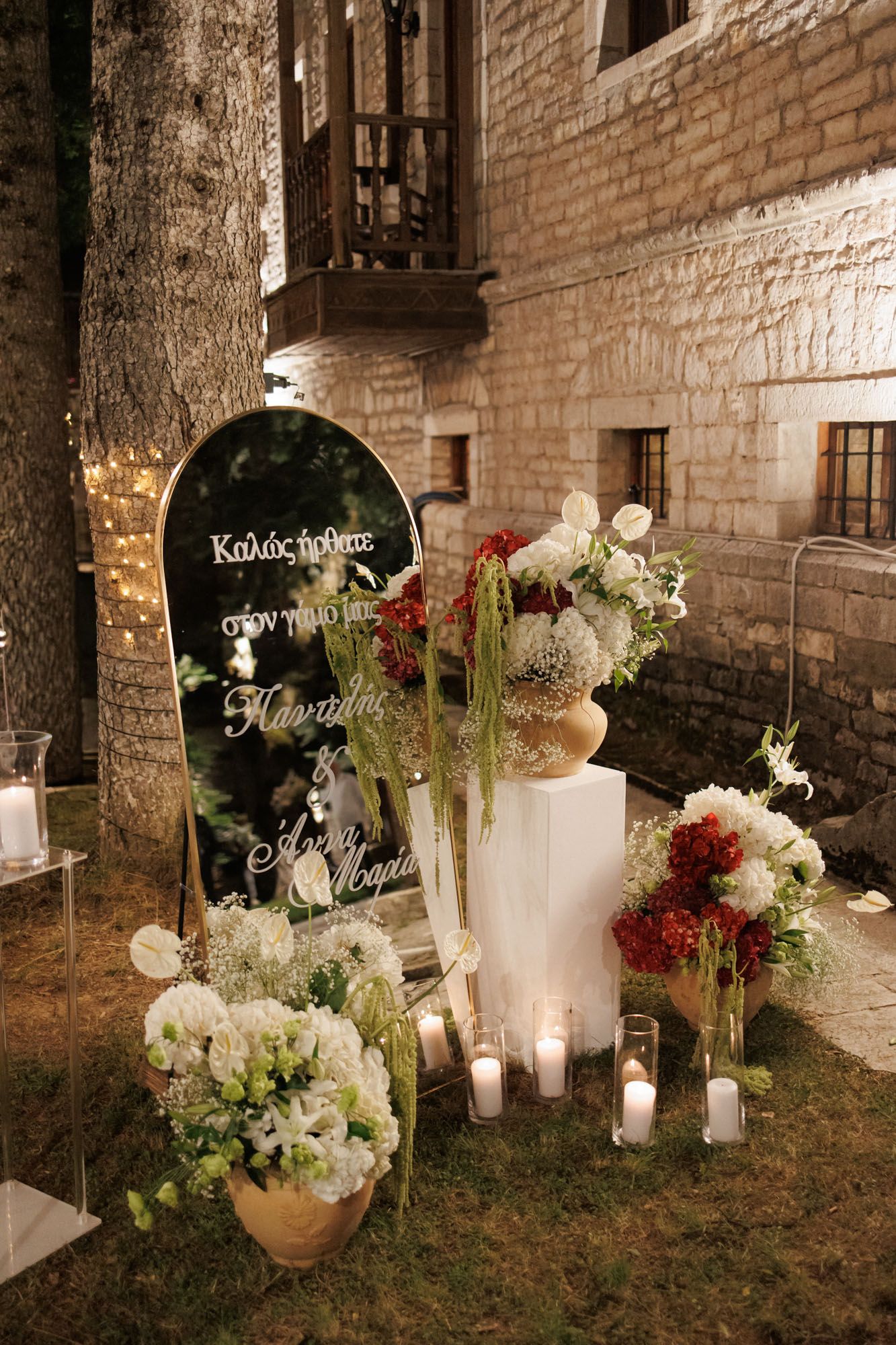 Elegant wedding welcome mirror sign surrounded by white and red floral arrangements and candles at night.