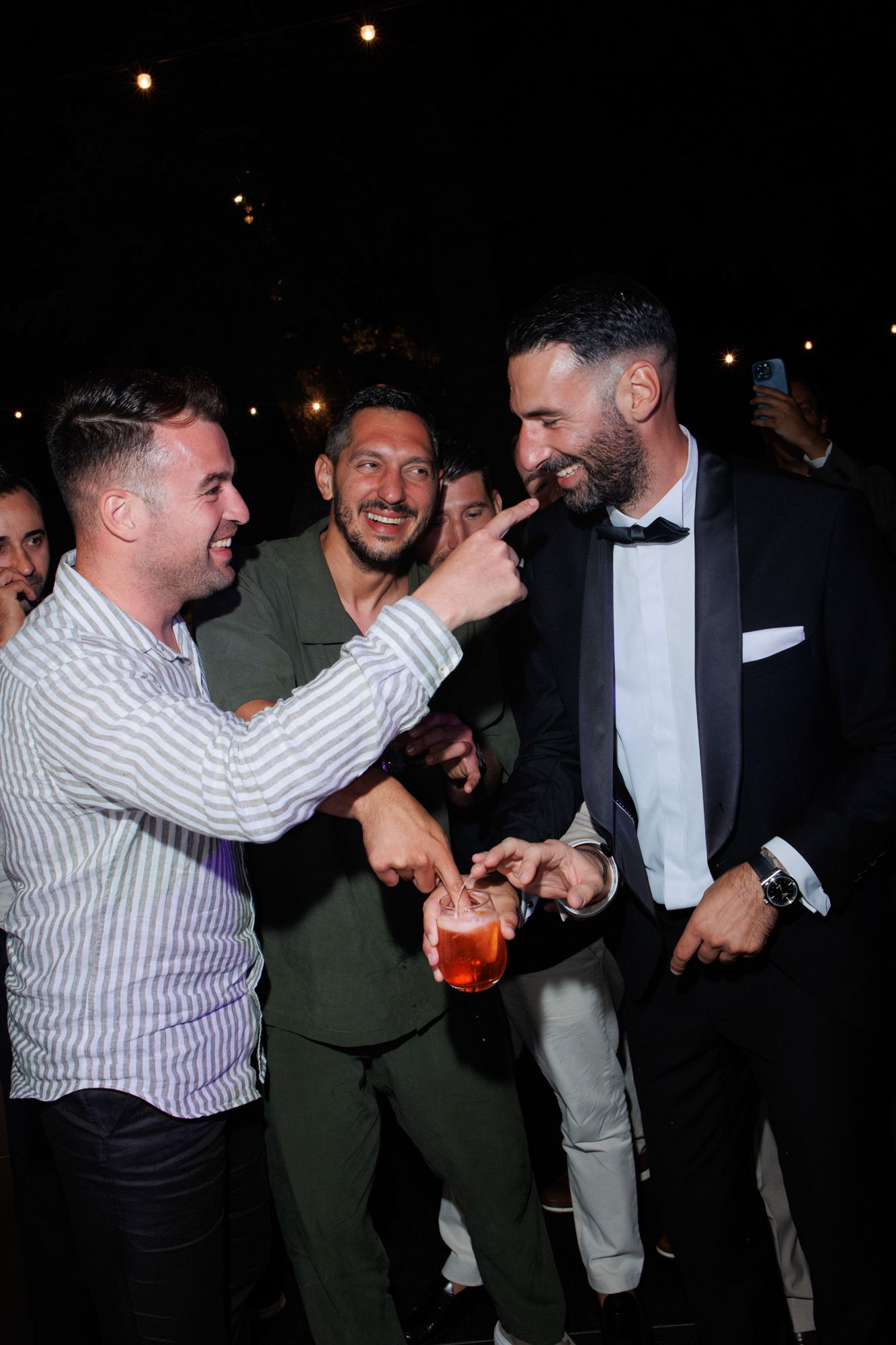 Groom in tuxedo laughing with guests at wedding reception party celebration at night.