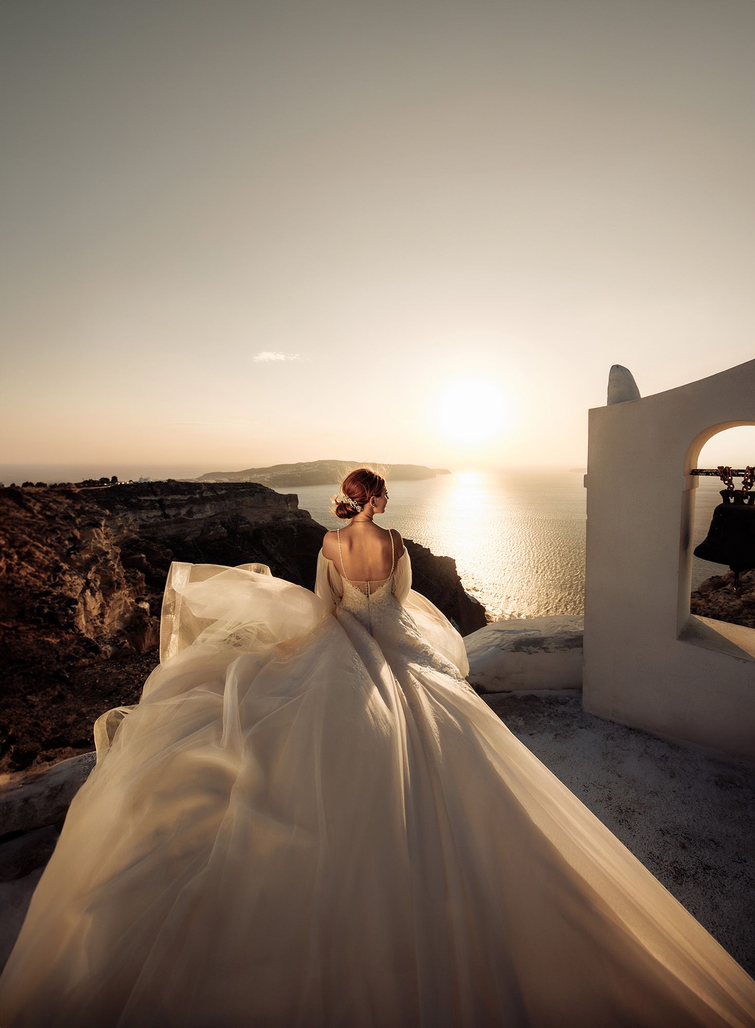 Bride in voluminous tulle wedding gown stands near white chapel bell tower at golden sunset in Santorini.