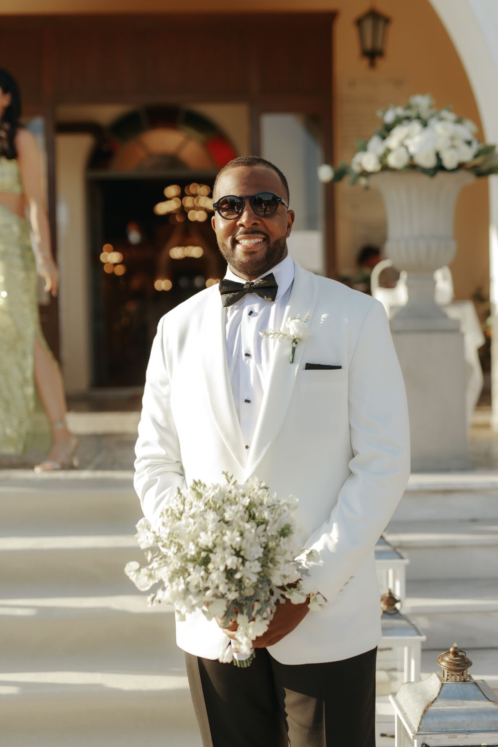 Smiling groom in white tuxedo jacket and black bow tie holding white floral bouquet outside wedding venue.