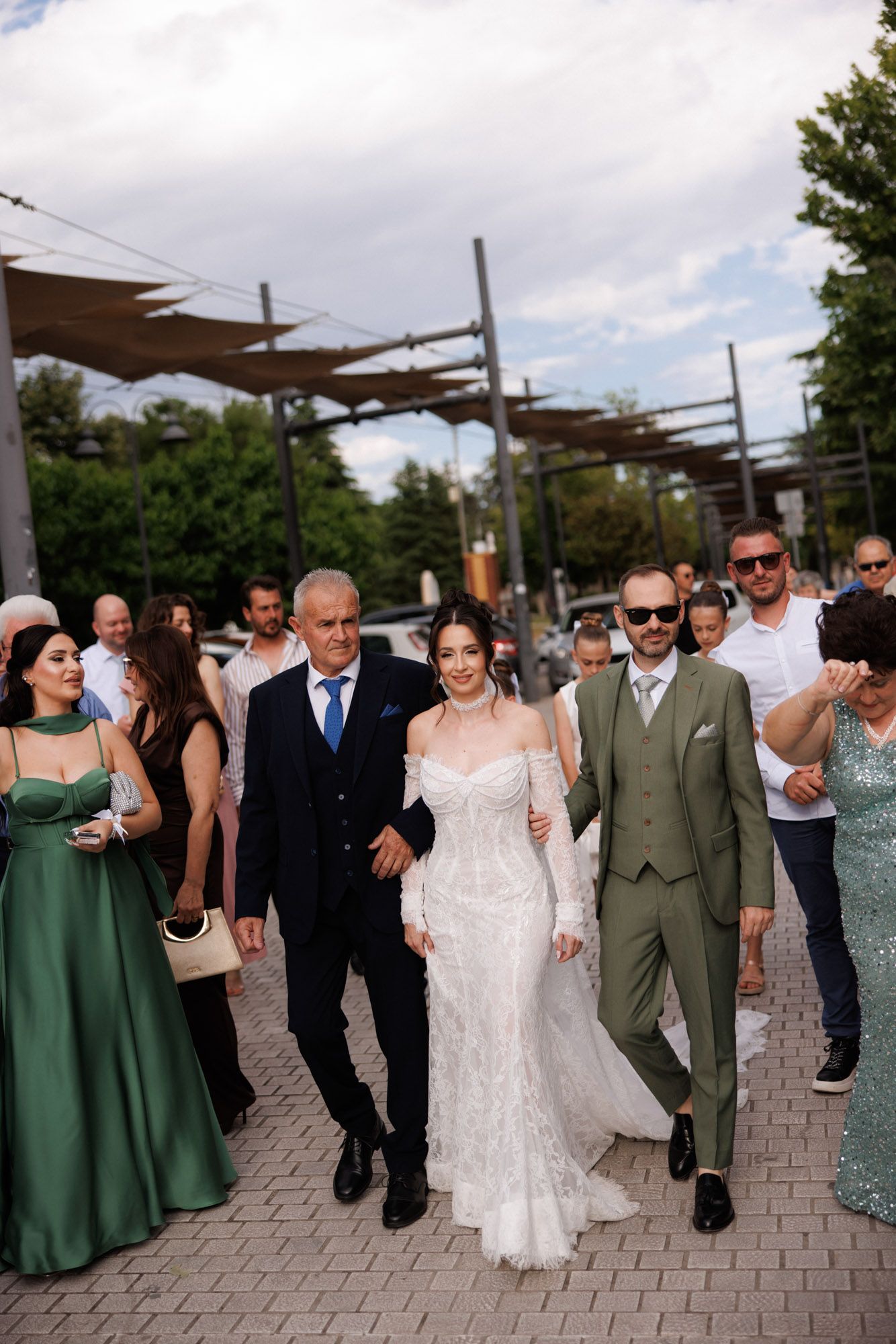 Bride in white lace gown walks with groom in green suit and older man, surrounded by guests in formal attire outdoors.