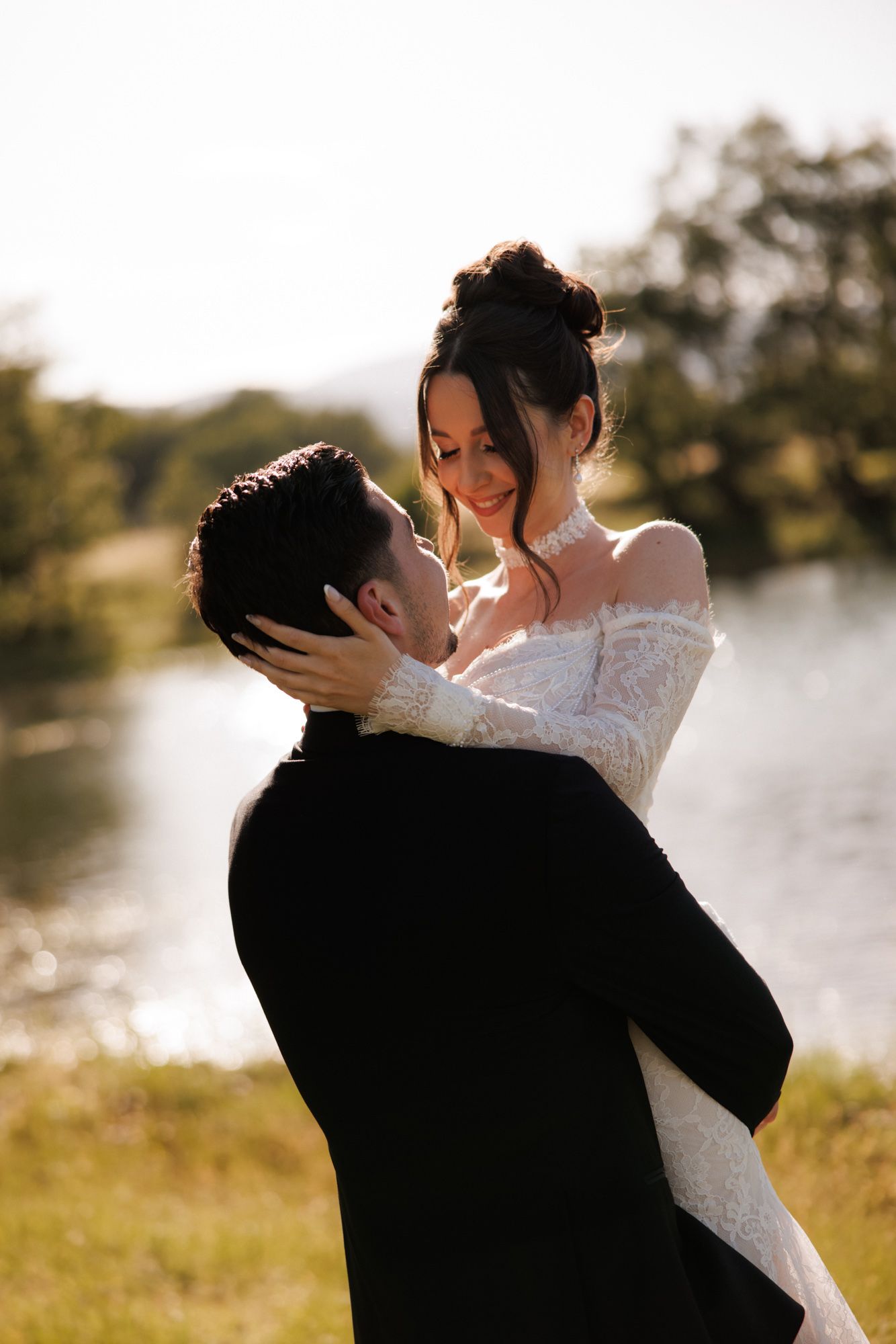 A bride in a lace off-shoulder gown embraces her groom in a black suit outdoors by a sunlit lake.