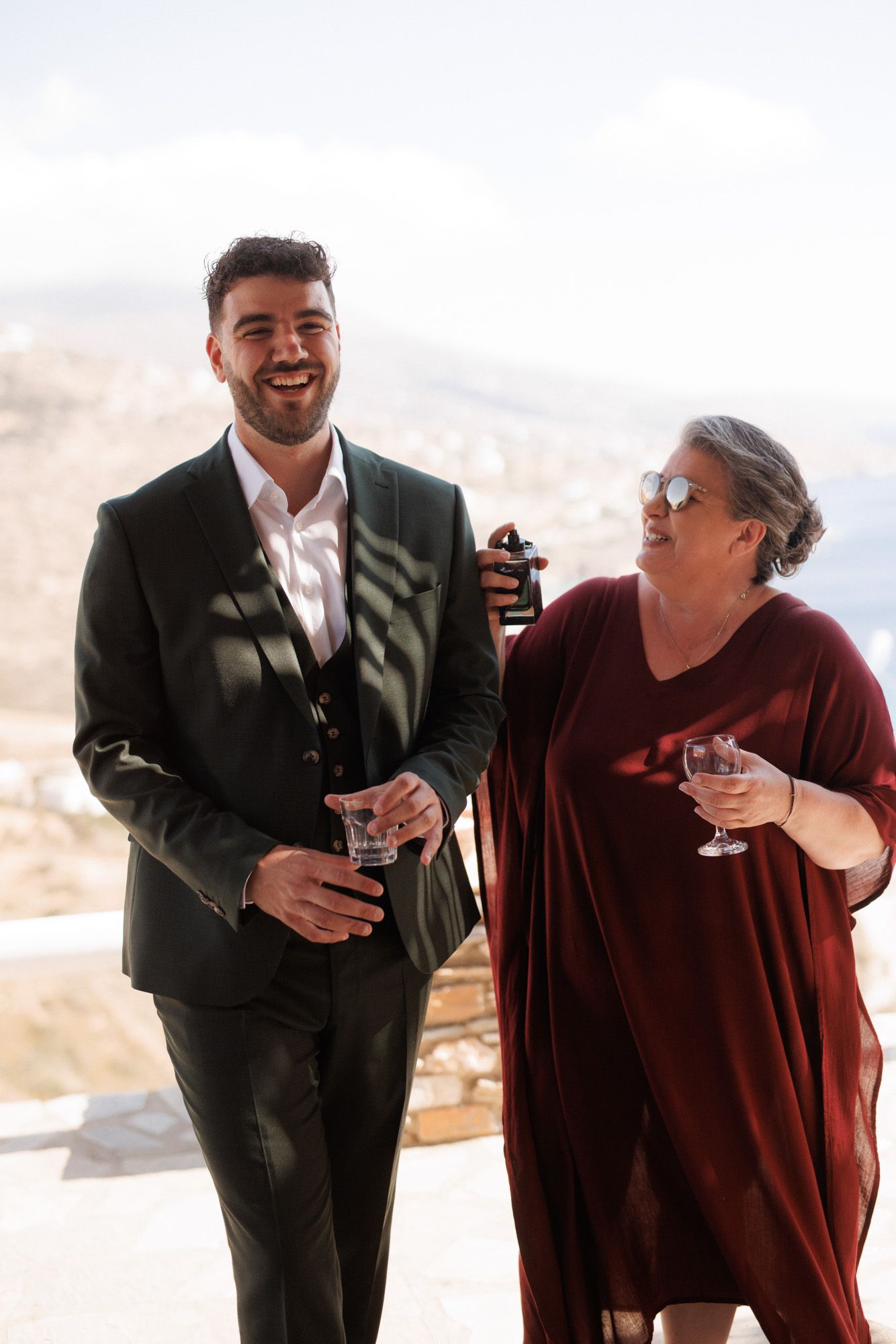 Smiling groom in dark suit with woman in burgundy dress enjoying drinks at Greek wedding.