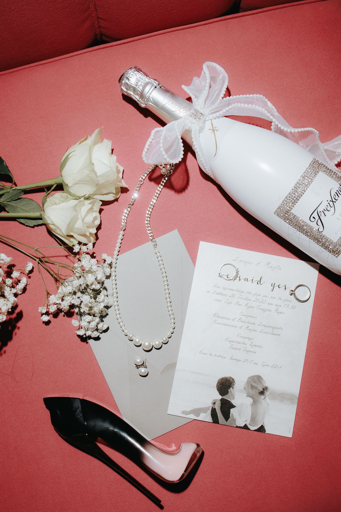 Elegant wedding flat lay with white champagne bottle, pearl necklace, roses, black heels, and bridal card on red surface.