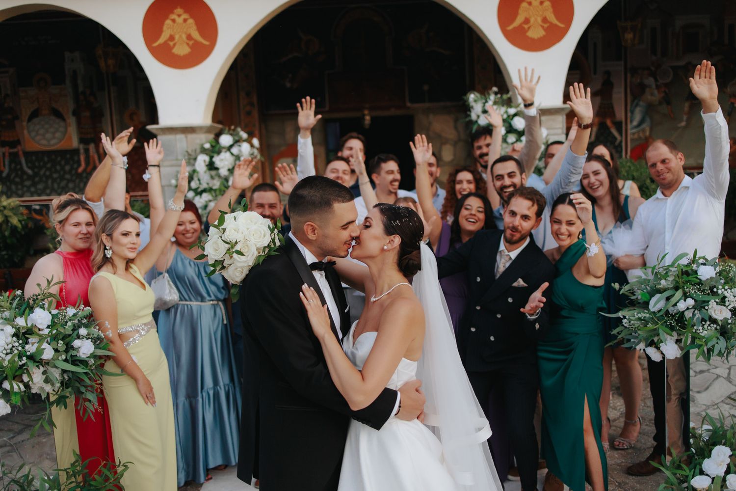 Bride and groom share a kiss surrounded by cheering wedding guests outside a historic church venue.