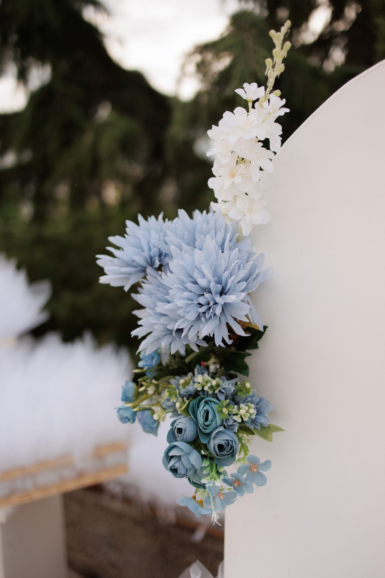 Blue and white floral arrangement with dahlias and roses on a white wedding arch, with green trees in the background.