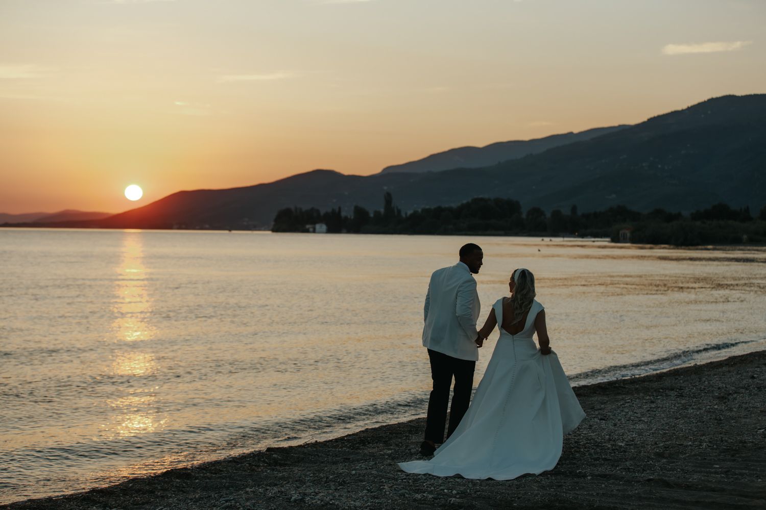Bride and groom holding hands on a beach at sunset with mountains and golden sky reflected on calm water.