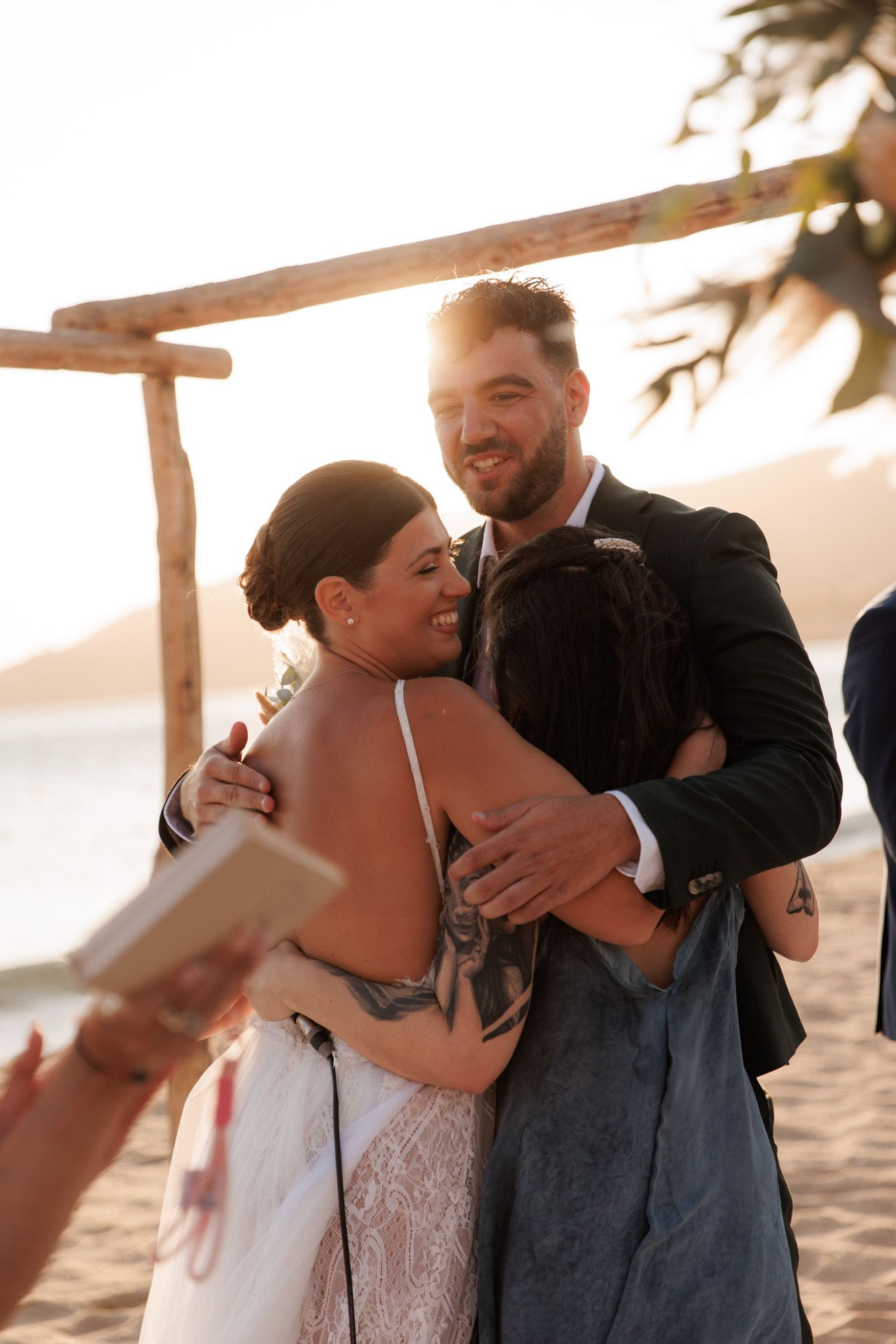 Bride and groom share a warm group hug with officiant at beachside wedding ceremony at sunset.